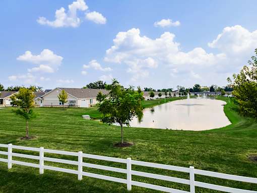 A white fence borders a grassy field with a pond in the distance.