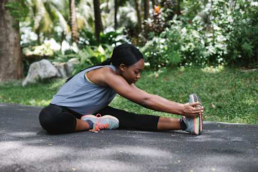 A woman stretches her leg while sitting on a mat outdoors.