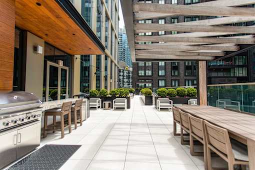 A patio with wooden furniture and a grill is surrounded by buildings.