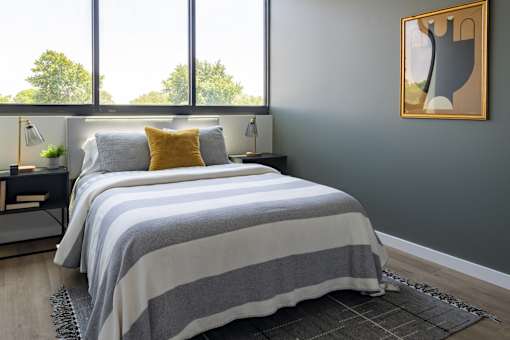 A bed with a grey and white striped comforter and a yellow pillow is in a room with a large window.