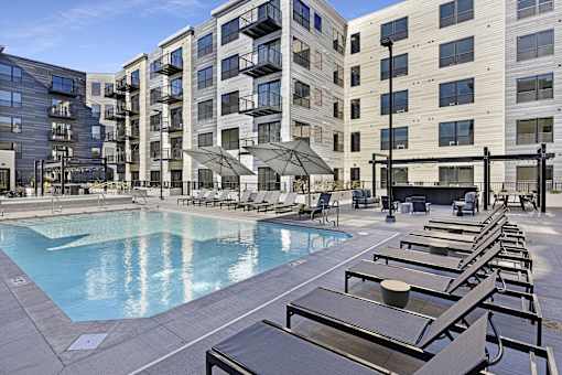 A large pool surrounded by sun loungers and umbrellas in front of apartment buildings.