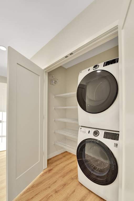 A white washing machine is in a small space between a wall and a shelf.