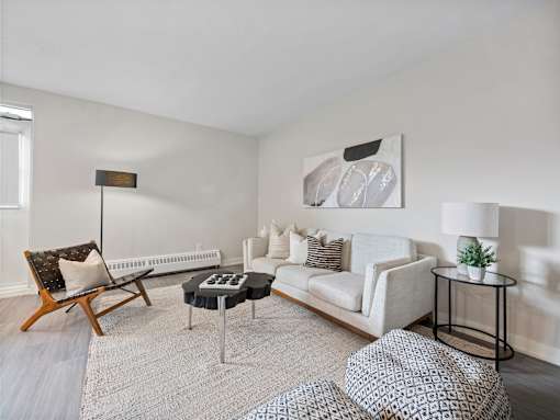 A living room with a white couch, a wooden chair, a black and white patterned ottoman, and a small table with a plant on it.