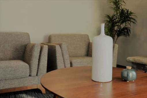 A white vase sits on a wooden table in front of a grey couch.