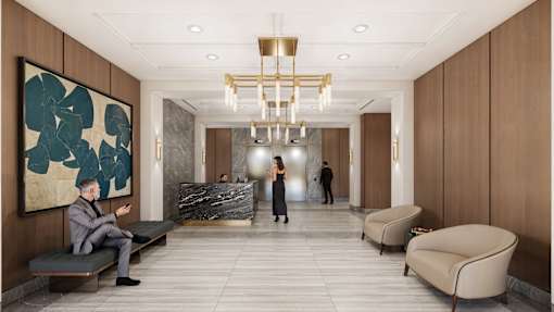 A man in a suit is sitting on a bench in a lobby with a woman walking in the background.
