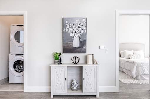 A white laundry room with a washer and dryer, a cabinet with a vase and plant on top, and a painting of flowers on the wall.