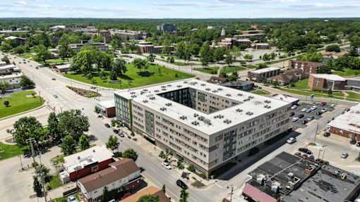 A large grey building is surrounded by smaller buildings and trees.