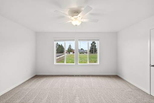 A room with a carpet floor, a ceiling fan, and a window showing a view of trees and a building.