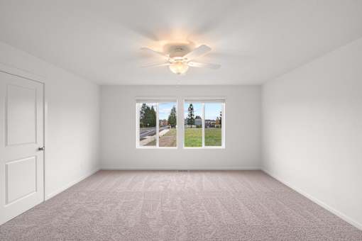 A room with a carpeted floor, a ceiling fan, and a window showing a view of trees and a road.