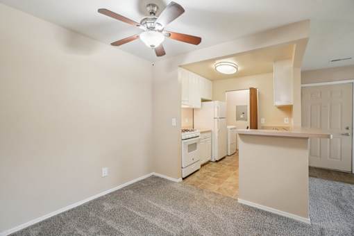 A room with a ceiling fan and a kitchen area in the background.