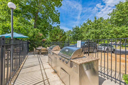 A barbecue grill is situated on a patio with a metal fence and trees in the background.