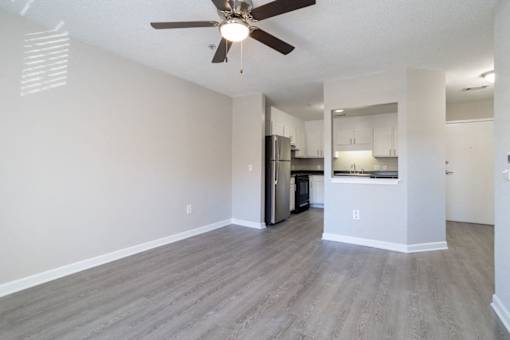 A spacious room with a ceiling fan and light fixture, hardwood flooring, and a kitchen area with stainless steel appliances.