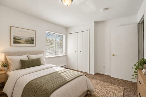 A bedroom with a bed, a nightstand, a window with blinds, a painting on the wall, a rug on the floor, and a plant on the nightstand.