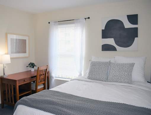 A bedroom with a bed, desk, chair, and a painting on the wall.