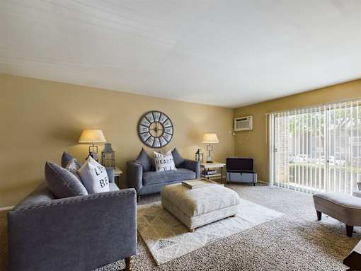A living room with a grey couch, a grey ottoman, a clock on the wall, and a window with blinds.