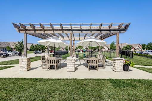A wooden pergola with two umbrellas provides shade for a picnic table and chairs.