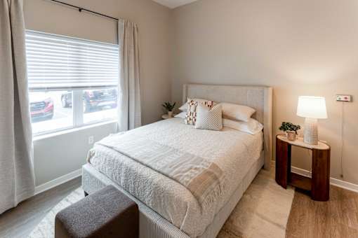 A bedroom with a large bed, a small table with a lamp, and a window with curtains.