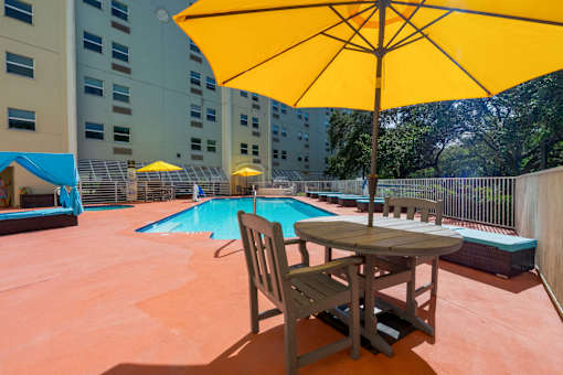 A yellow umbrella is on a table with a chair in front of a pool.