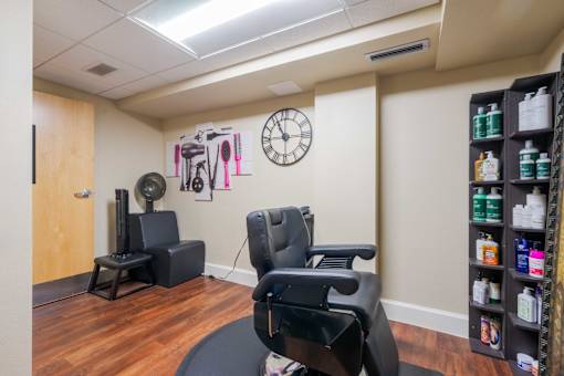 A room with a salon chair and a seated blow dryer in the background.  A clock and hair tool artwork is on the wall.