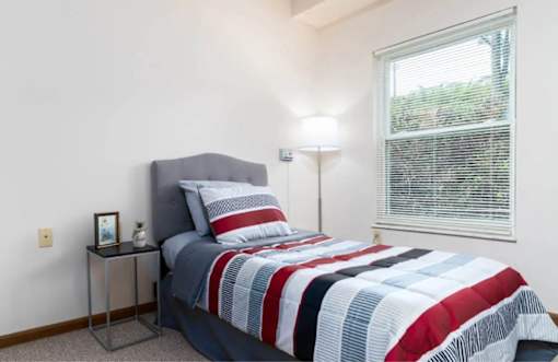 A bedroom with a bed, pillows, a lamp, and a small table with a picture frame on it.