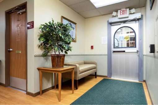 A room with a wooden door, a bench, a table with a plant on it, and a glass door with an exit sign above it.