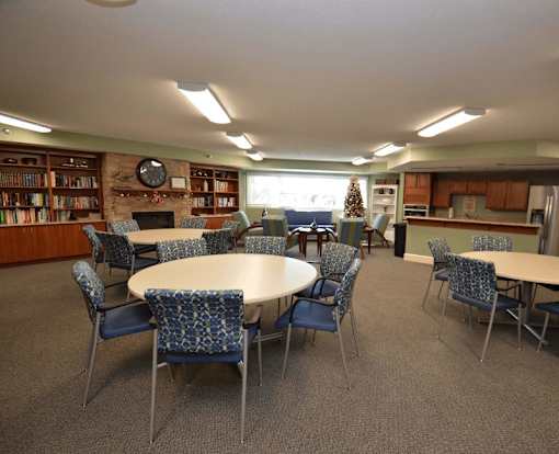 A large community room with round tables surrounded by chairs.  Bookshelves and a fireplace are in the background.