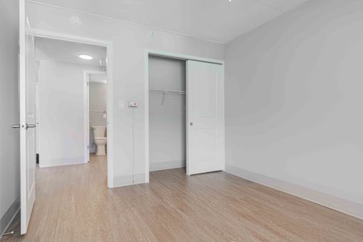 A bedroom with white walls, wood floors, double door closet and a view into the hallway and restroom.