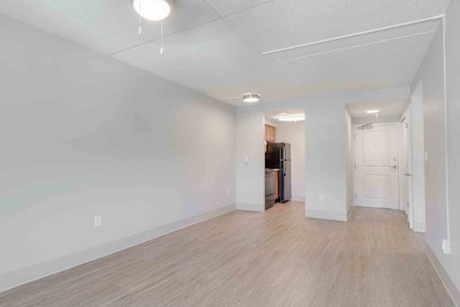 The living area with an overhead ceiling fan with light with white walls, wood floors looking into the kitchen and the main entry door.