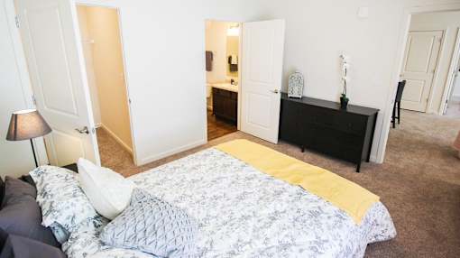 A bed with a yellow blanket is in the foreground of a room with a dresser and mirror.