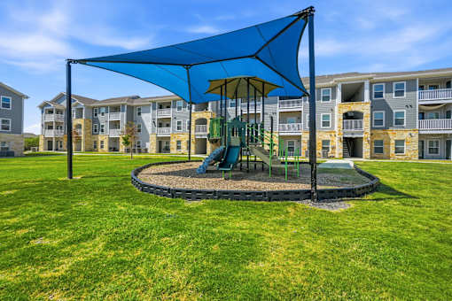 A playground with a blue canopy and a green slide is surrounded by apartment buildings.