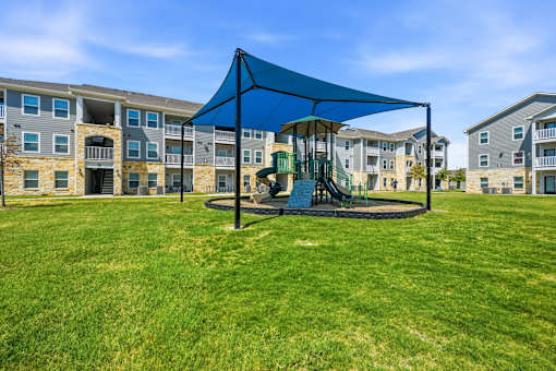 A playground with a blue canopy and a green slide is in front of apartment buildings.