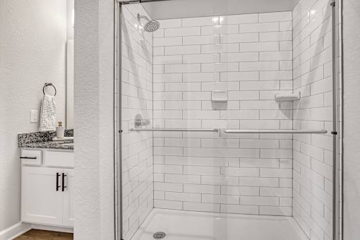 A white tiled shower with a glass door and a white vanity with a marble countertop.