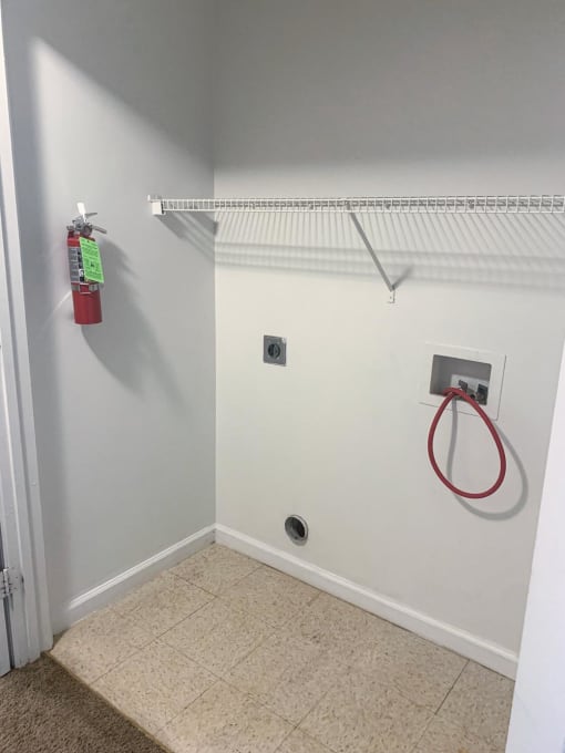 Small room with a white wall and a white shelf with a red fire extinguisher at Conner Court apartments in Connersville, IN