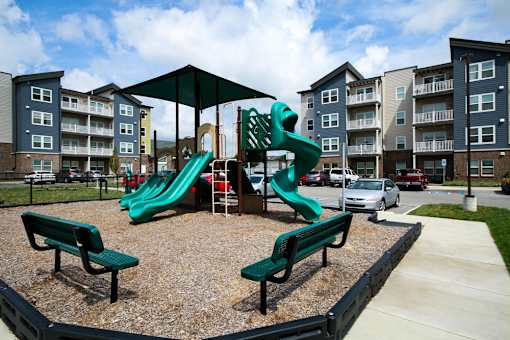 A playground with a green slide and two benches in front of apartment buildings.