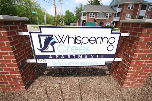 A sign for Whispering Creek Apartments is displayed in front of a brick wall.