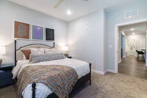 A bedroom with a bed, pillows, a blanket, a ceiling fan, and a chair in the hallway.