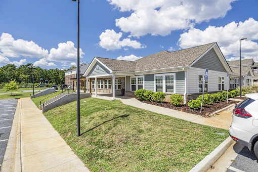 A building with a grey roof and white walls is surrounded by a grassy area and a parking lot.