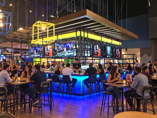 A group of people are sitting at tables in a restaurant with a bar in the background.