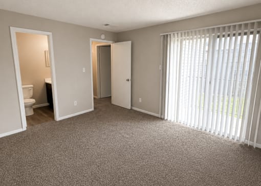 A room with a carpeted floor, a window with blinds, and a door leading to another room.