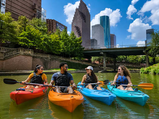 Four people kayaking on a river with a city skyline in the background.