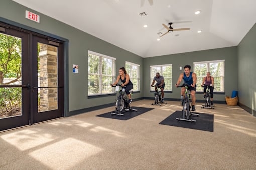A group of people are riding stationary bikes in a room with a ceiling fan.