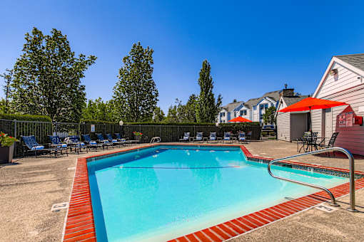 Happy Valley, OR, Apartments - Sunnyside Village - an Outdoor Gated Pool Area with Lounge Seating, Tables with Chairs and Umbrellas, Surrounding Trees and Shrubbery, and a View of the Property Building