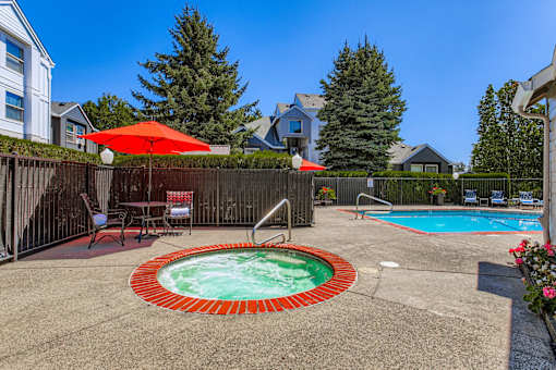 A hot tub sits in the middle of a backyard with a red umbrella and chairs around it.