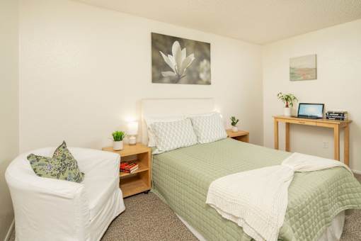 A bedroom with a bed, chair, and a small table with a laptop on it.