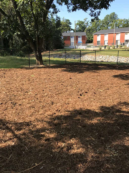 A dirt path with a fence and trees on the left and a building in the background.