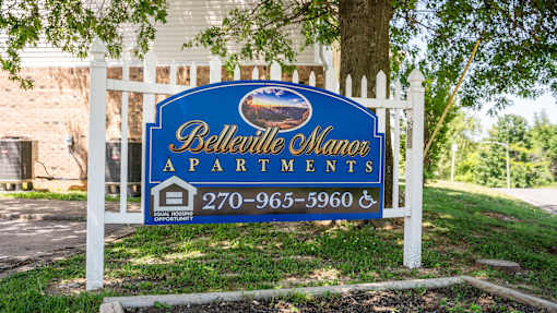 A sign for Belleville Manor Apartments is displayed in front of a white picket fence.
