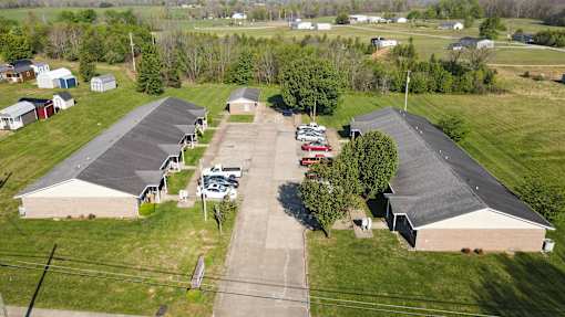 A parking lot is in the foreground of a grassy area with a building and a few cars.