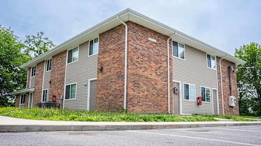 A brick apartment building with a grey roof and a parking lot in front.