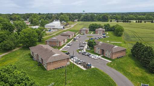 A large grassy area with a few buildings and cars parked in the lot.