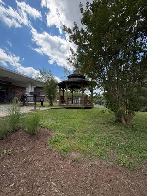 A gazebo sits in a grassy area with a tree and a house in the background.